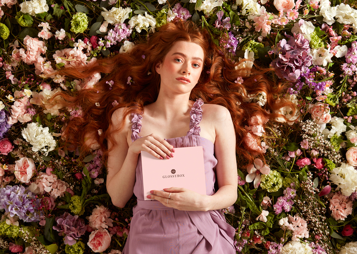 Red-haired woman in lilac dress lying on a bed of colorful flowers, holding a pink Glossybox beauty 